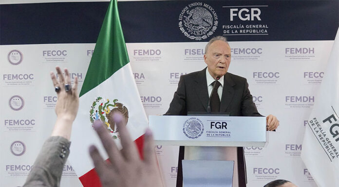Attorney General Alejandro Gertz Manero stands at a podium giving a report on the Jalisco extermination camp case, next to a Mexican flag as reporters raise their hands in the foreground