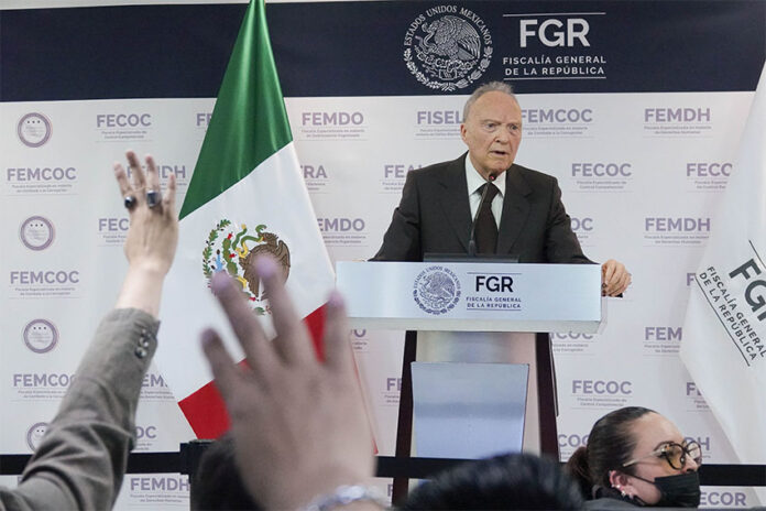 Attorney General Alejandro Gertz Manero stands at a podium giving a report on the Jalisco extermination camp case, next to a Mexican flag as reporters raise their hands in the foreground