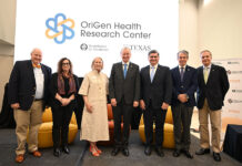 A group of older men and women stand in front of a screen reading OriGen Health Research Center