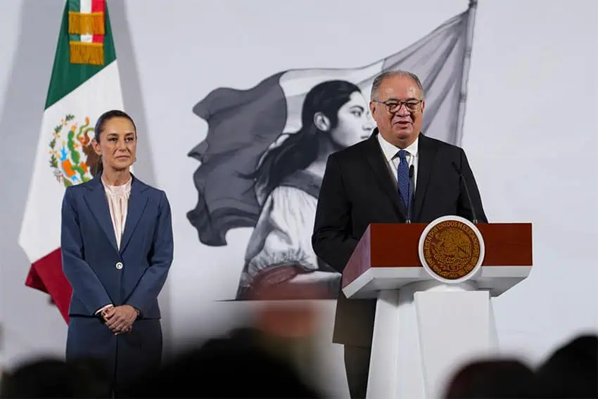 A stocky older man in glasses stands at a podium, as President Claudia Sheinbaum looks on