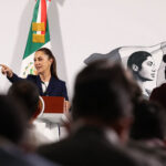 President Sheinbaum, visible at a podium behind a sea of silhouettes, points to the crowd to call on a reporter