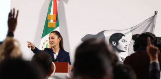 President Sheinbaum, visible at a podium behind a sea of silhouettes, points to the crowd to call on a reporter