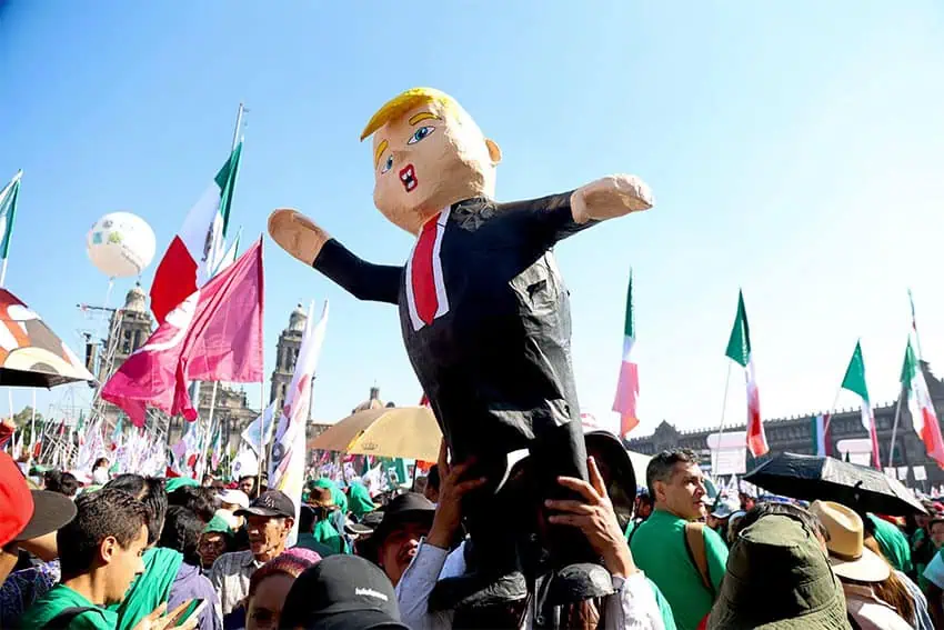 A member of the crowd carries an effigy of Trump in the Mexico City Zócalo while President Sheinbaum gives a speech on Trump's proposed tariffs, while other members of the crowd wave Mexican flags.