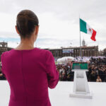 President Claudia Sheinbaum in a pink dress looks out over a crowd in Mexico City Zócalo with the National Palace and a Mexican flag in the background