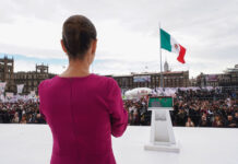 President Claudia Sheinbaum in a pink dress looks out over a crowd in Mexico City Zócalo with the National Palace and a Mexican flag in the background