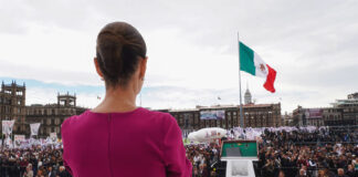 President Claudia Sheinbaum in a pink dress looks out over a crowd in Mexico City Zócalo with the National Palace and a Mexican flag in the background