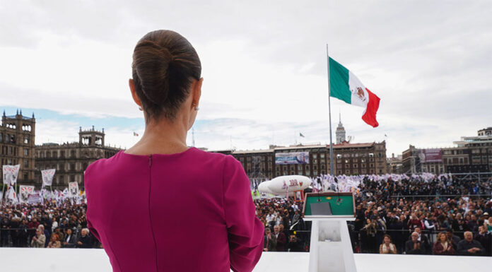 President Claudia Sheinbaum in a pink dress looks out over a crowd in Mexico City Zócalo with the National Palace and a Mexican flag in the background