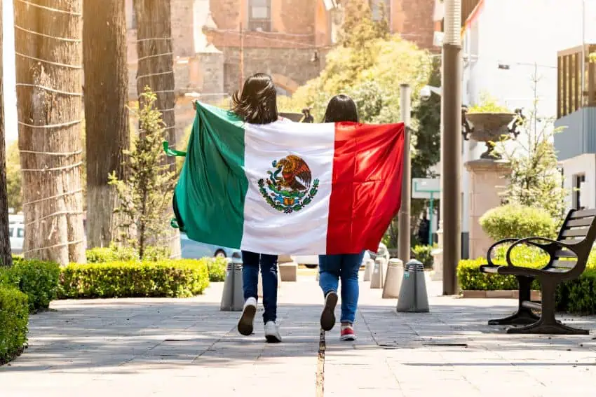 Two young Mexican women with their backs to the camera walking down a sidewalk in Mexico City holding the Mexican flag draped over their backs.