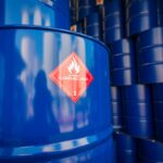 Blue barrels of oil stacked high in rows. One in front has a sticker saying "flammable liquid" on it.