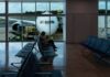 A passenger sitting in a chair at a gate in an empty section of an airport. In the background are large windows showing a plane parked on the runway, with its nose facing the window.