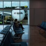 A passenger sitting in a chair at a gate in an empty section of an airport. In the background are large windows showing a plane parked on the runway, with its nose facing the window.