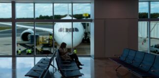 A passenger sitting in a chair at a gate in an empty section of an airport. In the background are large windows showing a plane parked on the runway, with its nose facing the window.