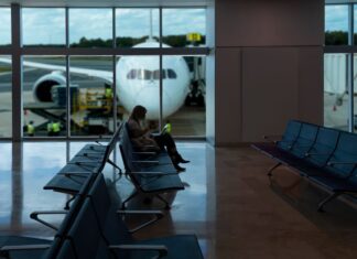 A passenger sitting in a chair at a gate in an empty section of an airport. In the background are large windows showing a plane parked on the runway, with its nose facing the window.