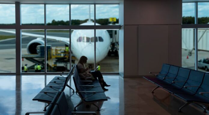 A passenger sitting in a chair at a gate in an empty section of an airport. In the background are large windows showing a plane parked on the runway, with its nose facing the window.