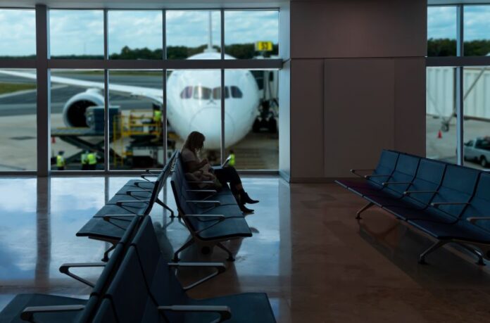 A passenger sitting in a chair at a gate in an empty section of an airport. In the background are large windows showing a plane parked on the runway, with its nose facing the window.