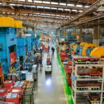 Workers laboring in the long aisle of a large factory floor with machines on either side towering over them.