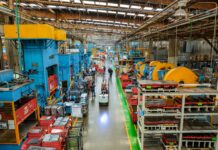 Workers laboring in the long aisle of a large factory floor with machines on either side towering over them.