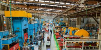 Workers laboring in the long aisle of a large factory floor with machines on either side towering over them.