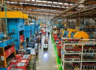 Workers laboring in the long aisle of a large factory floor with machines on either side towering over them.