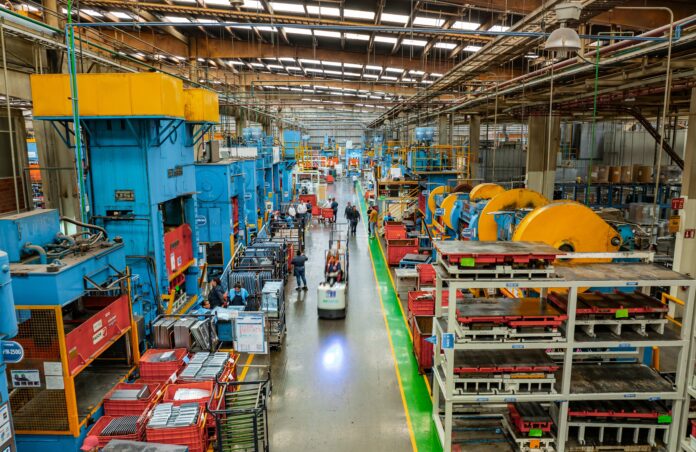 Workers laboring in the long aisle of a large factory floor with machines on either side towering over them.