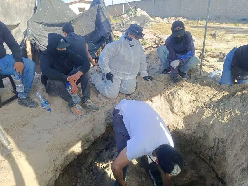 Several people in long sleeves and face masks gather around a hole in the ground where another person is digging