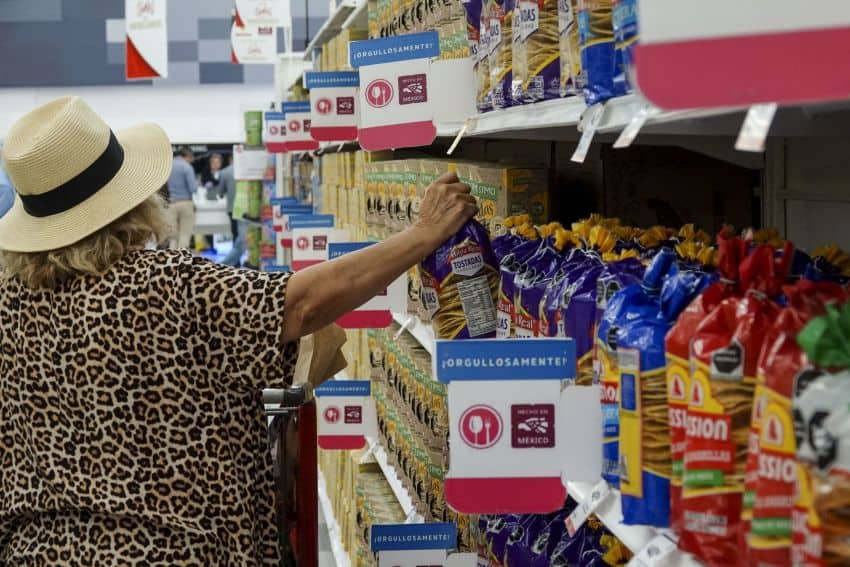 A Mexican woman with a leopard print shirt and a straw Panama style hat pushing a shopping cart through a supermarket aisle. She is selecting a bag of Mexican-made tostadas. Just behind her on the shelves is a shelf sign that says in Spanish "Proudly Made in Mexico" with a logo for the "Hecho en Mexico" program that looks like an ancient Aztec carving of an eagle