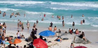 Tourists play on the sand and in the turquoise water of a Cancún beach, with small patches of sargassum seaweed visible between the umbrellas and beach towels