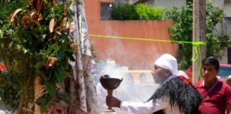 An Indigenous Mexican man in traditional dress holds a smoking large goblet in his right hand and offers it ritualistically to a shrine covered with vegetation and a large sheet of fabric with birds and plants printed on it. Behind him, a man in non-traditional clothing looks on with his eyes closed.