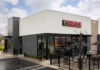 A concrete black and white Chiptole restaurant front, with the red Chipotle logo with white letters sitting on the building's top center. Outside, next to the front door is a small patio with tables for outdoor dining.