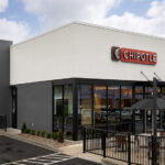 A concrete black and white Chiptole restaurant front, with the red Chipotle logo with white letters sitting on the building's top center. Outside, next to the front door is a small patio with tables for outdoor dining.