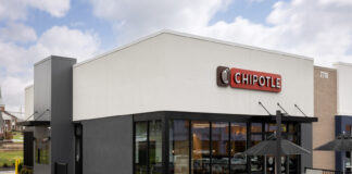A concrete black and white Chiptole restaurant front, with the red Chipotle logo with white letters sitting on the building's top center. Outside, next to the front door is a small patio with tables for outdoor dining.