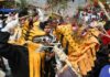 People dressed with paper mache masks made to look as jaguars. One person in costume is holding a crudely fashioned whip made of coiled rope and using it to lash another person in jaguar costume. A boy in normal clothing looks on