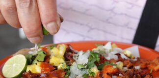 A plate of tacos al pastor being held in an unseen person's hand as they use their other hand to squeez lime juice onto the tacos