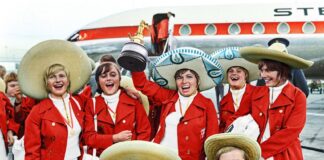 A team of women in Red jackets and sombreros lift the world cup trophy on the steps on a plane.