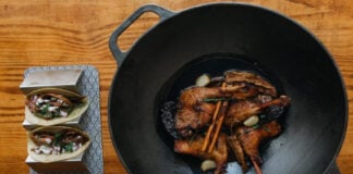 Duck legs cooked in a cast iron wok, with a taco serving dish and two tacos stuffed with meat and veggies on the side