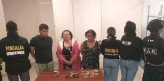 An elderly woman in a pink coat stands behind a table with a gun and packets of powder, next to two younger handcuffed people and several police agents