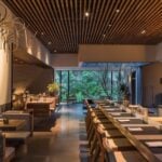 Japanese architecture influenced dining room at Pujol restaurant, with a long line of simple wood and thatched chairs in front of individual table in the center of the room and small two person booths of similar architecture along the left side of the room. At the far end of the photo is a floor-to-ceiling bay window with a view of a courtyard filled with lush green vegetation.
