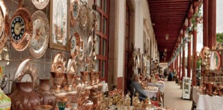 An artisan fair in a Mexican courtyard in a small town where hundreds of coppersmithed decorative objects are laid out on tables.