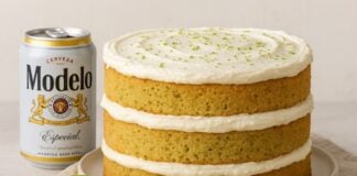 Image of a three-layer yellow cake with white frosting in between all the layers and on top, with grated lime zest on the top of the cake. The cake sits on a white plate, which is on a white tea towel. In front of the cake is half a lime and a tiny dipping bowl filled with honey. To screen left of the cake is an open can of Modelo Especial beer.