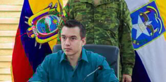 Ecuador President Daniel Noboa sits at a desk with a soldier in fatigues and an Ecuadorian flag behind him