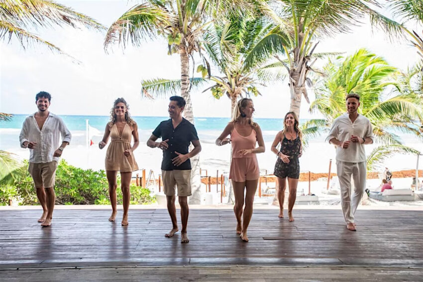 Six people in a line practice salsa on a wooden platform in front of a beach and palm trees