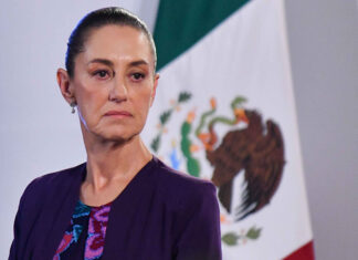 Claudia Sheinbaum with a serious expression and a Mexican flag