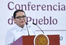 A dark-haired woman speaks at a podium emblazoned with the emblem of the Mexican government.