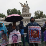 Protesters hold signs showing the faces of the disappeared on a rainy day in front of a Mexico City landmark