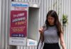 A Young woman in a gray sleeveless top and a black skirt and carrying a white messenger bag over her shoulder walks past a telephone booth in Mexico City that has a sign urging Mexico City residents to vote in the upcoming judicial election on June 1, 2025.