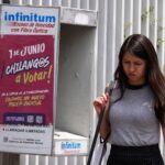 A Young woman in a gray sleeveless top and a black skirt and carrying a white messenger bag over her shoulder walks past a telephone booth in Mexico City that has a sign urging Mexico City residents to vote in the upcoming judicial election on June 1, 2025.