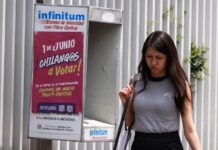 A Young woman in a gray sleeveless top and a black skirt and carrying a white messenger bag over her shoulder walks past a telephone booth in Mexico City that has a sign urging Mexico City residents to vote in the upcoming judicial election on June 1, 2025.