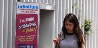 A Young woman in a gray sleeveless top and a black skirt and carrying a white messenger bag over her shoulder walks past a telephone booth in Mexico City that has a sign urging Mexico City residents to vote in the upcoming judicial election on June 1, 2025.