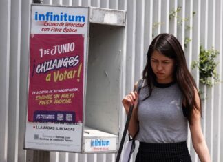 A Young woman in a gray sleeveless top and a black skirt and carrying a white messenger bag over her shoulder walks past a telephone booth in Mexico City that has a sign urging Mexico City residents to vote in the upcoming judicial election on June 1, 2025.