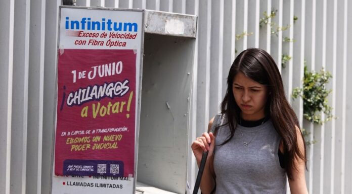 A Young woman in a gray sleeveless top and a black skirt and carrying a white messenger bag over her shoulder walks past a telephone booth in Mexico City that has a sign urging Mexico City residents to vote in the upcoming judicial election on June 1, 2025.